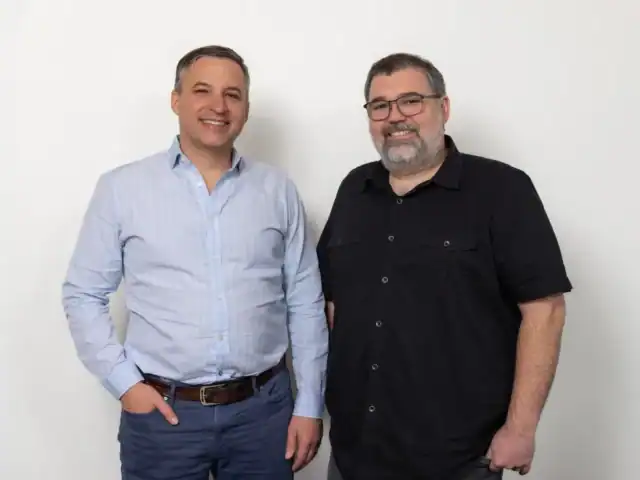Two men stand side by side against a plain white background, one in a light blue button-up shirt and the other in black, reflecting the modern style of AI employees at a fast-paced startup.
